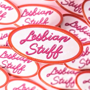May include: Oval stickers with the words "Lesbian Stuff" in pink script. The stickers have a white background and a red border. Several stickers are arranged on a pink surface.