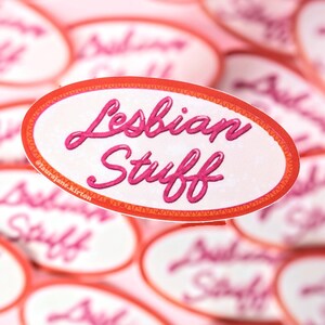 May include: Oval stickers with the words "Lesbian Stuff" in pink script, outlined in a red border. The stickers are scattered on a pink background, with some in focus and others blurred.