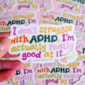 May include: A die-cut vinyl sticker featuring the text I don't struggle with ADHD, I'm actually really good at it. in a multi-colored, wavy, retro-style font. The design includes a small yellow smiley face and an orange heart outline.