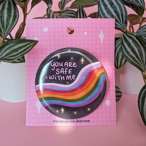 May include: A black and white button with a rainbow arc and the text "You are safe with me" and the social media handle "@laurajane.kirton" on a pink background.