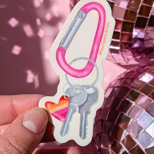 May include: A die-cut sticker featuring a bright pink carabiner attached to a silver metal keyring with two silver keys and a small heart-shaped charm. The heart charm displays horizontal stripes in shades of orange, pink, and red. The text @laurajane.kirlon is printed in small black letters along the side of the carabiner.
