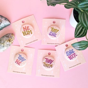May include: Five round buttons with different pronoun sets on each. The buttons are white with a glittery background and colorful text. The text on the buttons reads "He Him", "They She", "They He", "They Them", and "She Her".