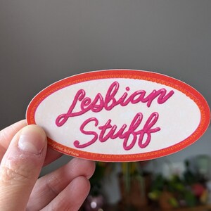 May include: An oval sticker with the words "Lesbian Stuff" in pink cursive lettering. The sticker has a white background with an orange and red border. The sticker is held by a hand.