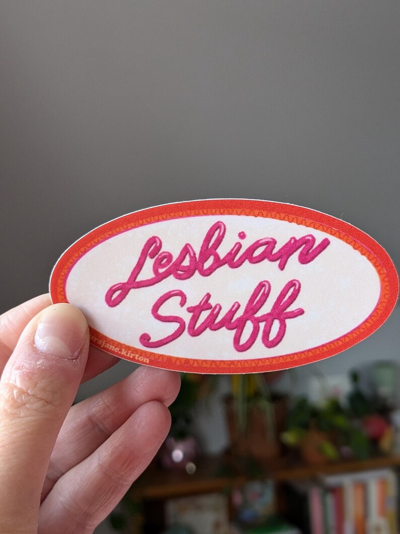 May include: An oval sticker with the words "Lesbian Stuff" in pink cursive lettering. The sticker has a white background with an orange and red border. The sticker is held by a hand.