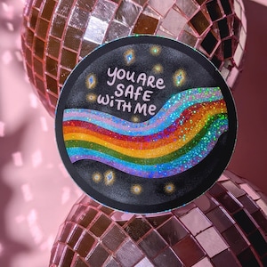 May include: A circular sticker with a black background featuring a glittery, multi-colored rainbow wave across the center. The text "YOU ARE SAFE WITH ME" is written in white, hand-lettered font above the rainbow, surrounded by small, sparkling gold stars.