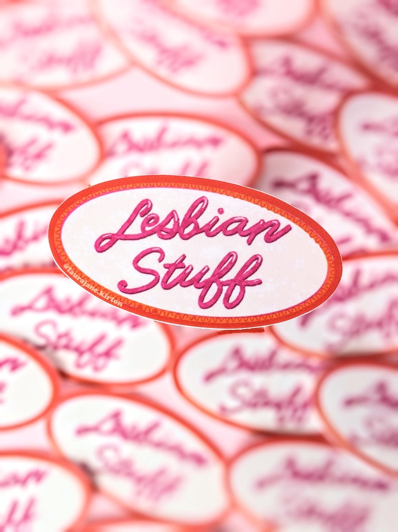 May include: Oval stickers with the words "Lesbian Stuff" in pink script, outlined in a red border. The stickers are scattered on a pink background, with some in focus and others blurred.
