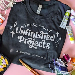 Op de afbeelding: Een zwart t-shirt met de witte tekst "The Society of Unfinished Projects" en de zin "So many ideas. So little focus". Knutselspullen, waaronder verftubes, lijm en borduurgaren, omringen het shirt op een roze ondergrond.