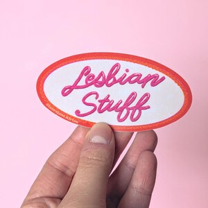 May include: An oval sticker with the words "Lesbian Stuff" in pink script. The sticker has a white background with a red and orange border. The sticker is held against a pink background.