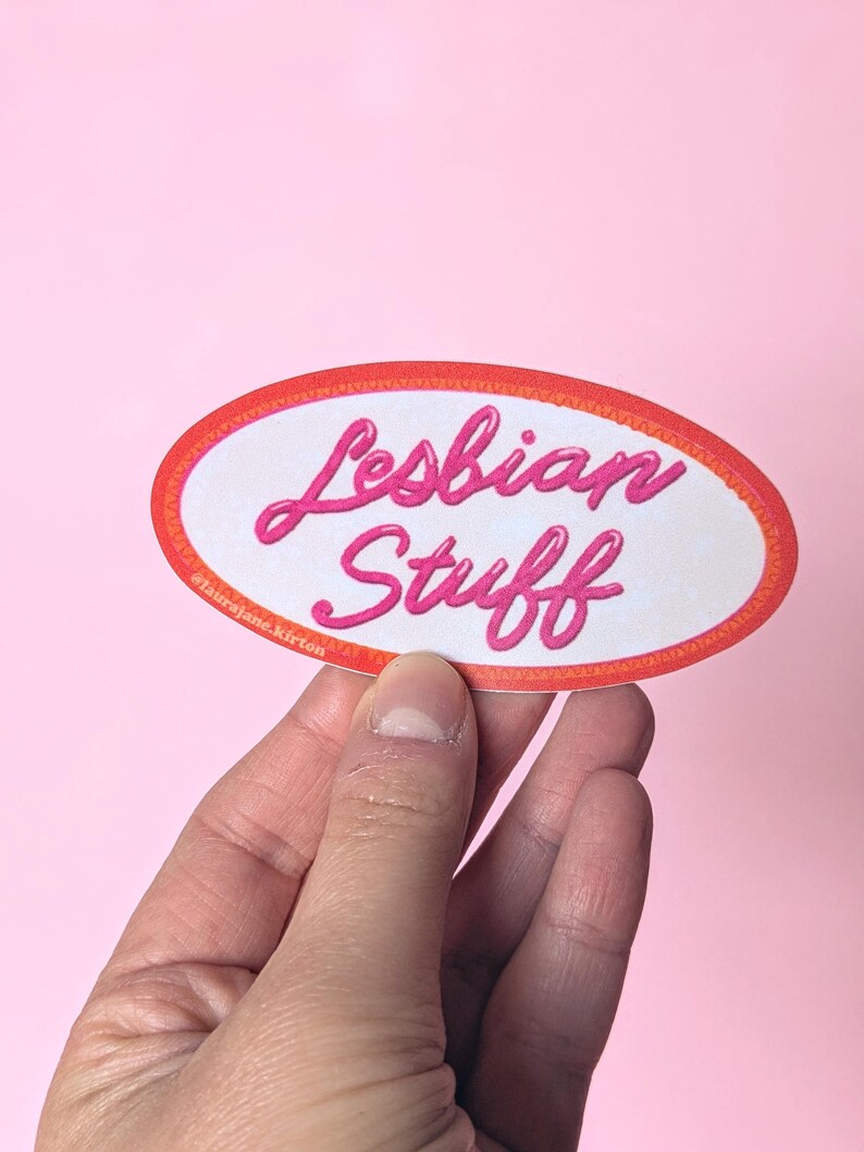 May include: An oval sticker with the words "Lesbian Stuff" in pink script. The sticker has a white background with a red and orange border. The sticker is held against a pink background.