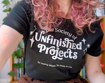 May include: A black short-sleeved t-shirt featuring white text that reads The Society of Unfinished Projects and So many ideas. So little focus, accented with white star graphics. The shirt is paired with brown trousers featuring a tie-waist detail.