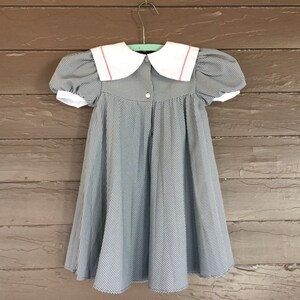 May include: A black and white gingham dress with a white Peter Pan collar and red trim. The dress has puffed sleeves and a full skirt.