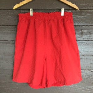 May include: Bright red shorts with an elastic waistband, hanging on a wooden hanger. The shorts are made of a smooth fabric and appear to be lightweight. The shorts are a solid color and have a simple, classic design.