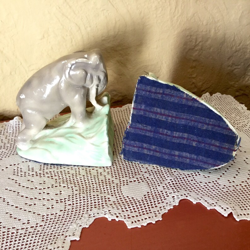 May include: A pair of ceramic bookends featuring a gray elephant standing on a green base. The other bookend is a plain, gray wedge with a blue fabric lining.