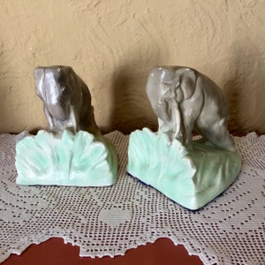 May include: A pair of gray ceramic elephant bookends with green leafy bases. The elephants are facing inward and have their trunks raised.
