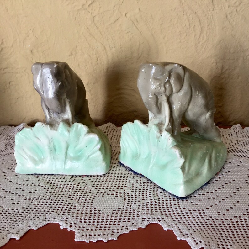 May include: A pair of gray ceramic elephant bookends with green leafy bases. The elephants are facing inward and have their trunks raised.