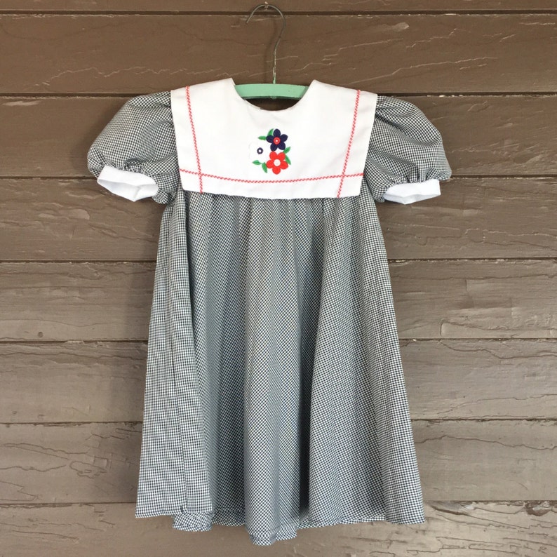 May include: A black and white gingham dress with a white Peter Pan collar and red trim. The collar has a colorful embroidered flower design.