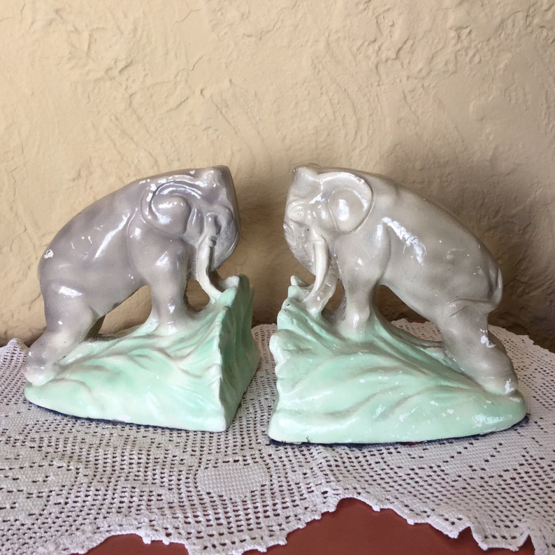 May include: A pair of vintage elephant bookends made of plaster. The elephants are gray and have green bases. The bookends are in good condition.