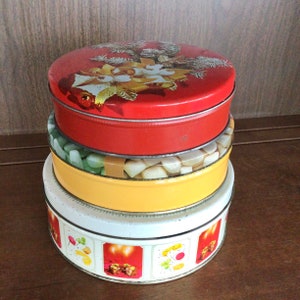 May include: A stack of four round vintage tins. The top tin is red with a Christmas design. The second tin is green and yellow, filled with candies. The third tin is yellow. The bottom tin is white with red and gold illustrations.
