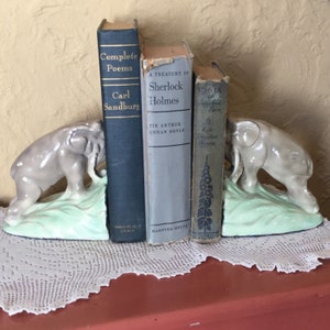 May include: A pair of gray elephant bookends with green bases holding up three books. The books are "Complete Poems" by Carl Sandburg, "A Treasury of Sherlock Holmes" by Sir Arthur Conan Doyle, and "Rebecca of Sunnybrook Farm" by Kate Douglas Wiggin.