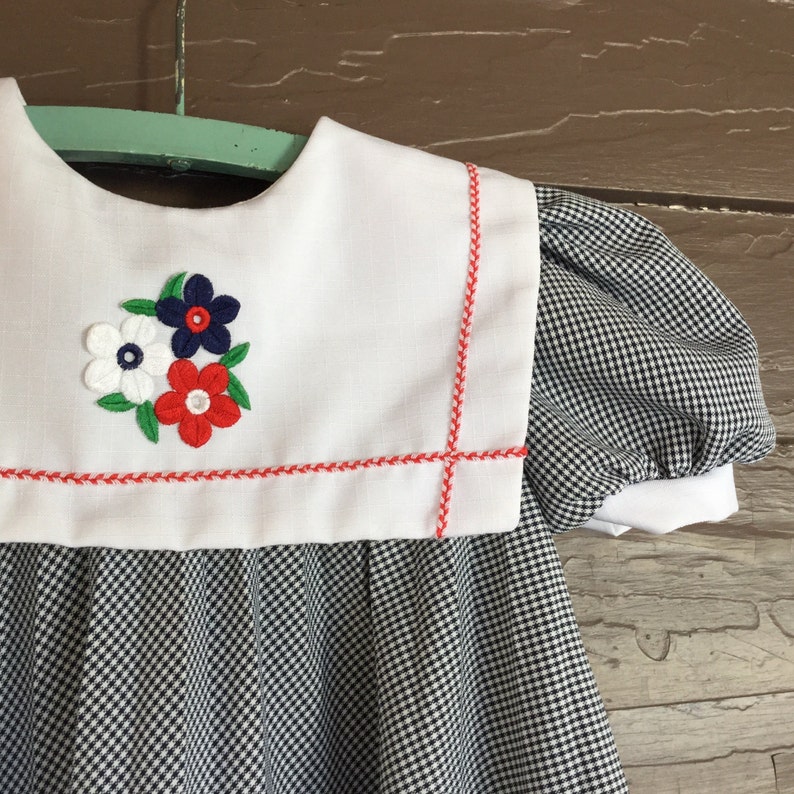 May include: A white dress with a gingham pattern in black and white. The dress has a white collar with red stitching and a red, white, and blue embroidered flower design.