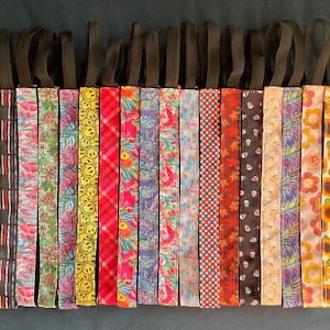 May include: A collection of colorful headbands with various patterns, including stripes, smiley faces, floral designs, and skulls. Each headband has a black elastic band for a secure fit. The headbands are displayed against a dark background.