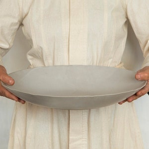 Puede incluir: Un gran bol de servir redondo de cerámica gris claro sostenido por dos manos.