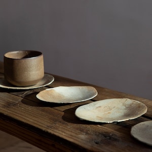 May include: A set of rustic ceramic teacups, saucers, and small serving dishes. The teacup and saucer are brown, while the dishes are cream and brown. The items are arranged on a weathered wooden surface, creating a natural, earthy aesthetic.