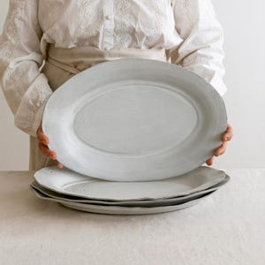 May include: A stack of three oval-shaped, off-white ceramic platters. The top platter is held up, revealing the other two below. The platters have a slightly textured surface and a simple, elegant design, suitable for serving food.