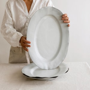 Puede incluir: Platos de servir ovalados blancos con una superficie texturizada y un borde oscuro. Un plato se sostiene sobre otro. Los platos son sostenidos por una persona que lleva una camisa blanca y un delantal. Los platos están sobre un mantel blanco.
