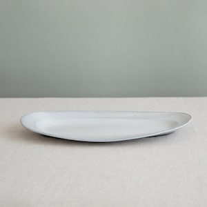 Pode incluir: Uma travessa de servir de cerâmica branca em forma oval. A travessa tem uma borda ligeiramente elevada e um acabamento fosco. A superfície tem salpicos subtis. O fundo é de cor neutra.