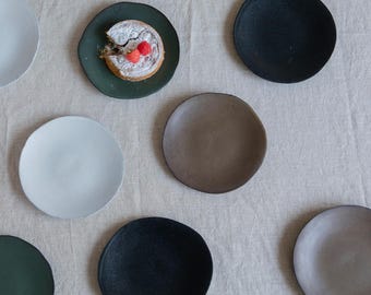 May include: A collection of nine ceramic plates in various colours and shapes, including green, black, brown, and white. The plates are arranged on a white linen surface. One plate features a small pastry with red berries.