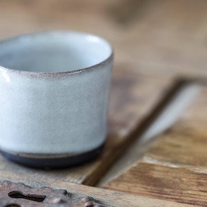 May include: A small, white ceramic cup with a dark grey rim. The cup is sitting on a wooden surface.
