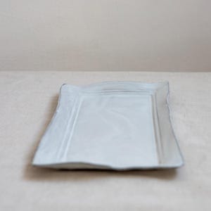 Puede incluir: Una fuente rectangular de cerámica blanquecina con un borde ligeramente irregular y texturizado. La fuente tiene un centro empotrado y un diseño sutil y minimalista, adecuado para servir aperitivos o platos pequeños.