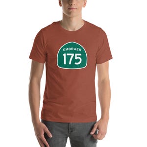 May include: A short-sleeved, rust-colored t-shirt featuring a green and white graphic. The graphic includes the word "EMBRAER" above the number "175" inside a rounded rectangle.
