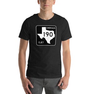 May include: Dark gray t-shirt featuring a white graphic of the Texas state outline with the text "EMBRAER 190 EJET" inside a white square. The shirt is short-sleeved and has a crew neck.