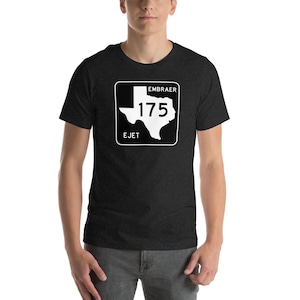 May include: Dark gray t-shirt featuring a white graphic of a Texas road sign with the text "EMBRAER 175 EJET". The graphic is a stylized outline of the state of Texas with the number 175 inside. The shirt is short-sleeved and has a crew neck.