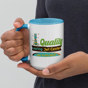 May include: A white ceramic mug with a bright blue interior and handle. The exterior features a graphic of a control tower and the text Quality Learing Jet Center, with the tagline Lear to Fly Here! printed in green, brown, and orange colours.