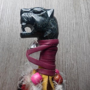 May include: A dark green carved stone wand featuring a jaguar head. The wand is wrapped in burgundy leather and adorned with a clear crystal, pink beads, yellow leather, feathers, and red accents. The wand is a unique spiritual item.