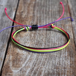 May include: A multi-strand cord bracelet featuring neon yellow, hot pink, black, and purple strings. The bracelet includes an adjustable sliding knot closure with two long, loose cord ends in red and purple, secured by a small black barrel bead.