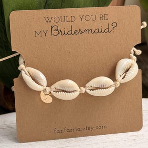 May include: A beige cord bracelet featuring four white cowrie shells and a small gold-toned circular charm with the letter H. The bracelet is displayed on a rectangular brown card with the text WOULD YOU BE MY Bridesmaid? and fanfarria.etsy.com.