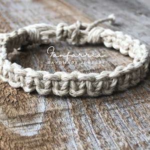 May include: A beige macrame bracelet crafted from natural fiber cord, featuring a repeating knotted pattern and an adjustable tie closure. The text Fanfarria HANDMADE JEWELRY is printed in white across the center of the bracelet.