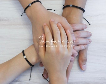 May include: Four hands wearing black friendship bracelets with silver heart charms. The bracelets are tied together, symbolizing a bond of friendship. The text "Fanfarria Handmade Jewelry" is visible in the image.