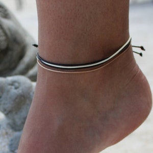 May include: Ankle bracelets in black, white, and gold. The bracelets are thin and delicate, with a simple design. The image shows the bracelets on an ankle, with a blurred background of a statue and sand.