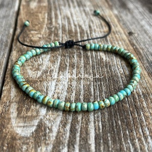 May include: A turquoise blue beaded bracelet with a black cord closure. The bracelet is made of small, round beads with a speckled pattern.