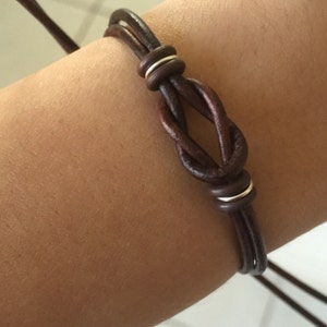 May include: A dark brown leather bracelet featuring a knot design with silver-colored metal accents. The bracelet is made of multiple strands of leather and is designed to be adjustable. The bracelet is shown on an arm.