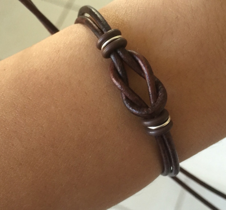 May include: A dark brown leather bracelet featuring a knot design with silver-colored metal accents. The bracelet is made of multiple strands of leather and is designed to be adjustable. The bracelet is shown on an arm.