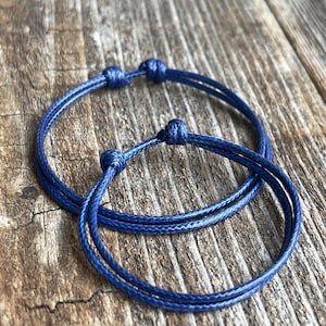 May include: Two blue friendship bracelets made of thin cord, tied with knots. The bracelets are on a wooden surface.