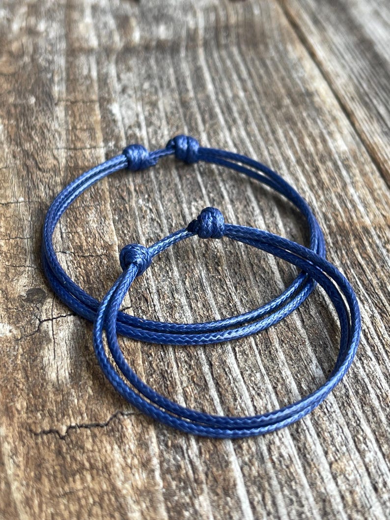 May include: Two blue friendship bracelets made of thin cord, tied with knots. The bracelets are on a wooden surface.