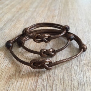 May include: Three brown cord bracelets with knotted ends. The bracelets are stacked on top of each other.
