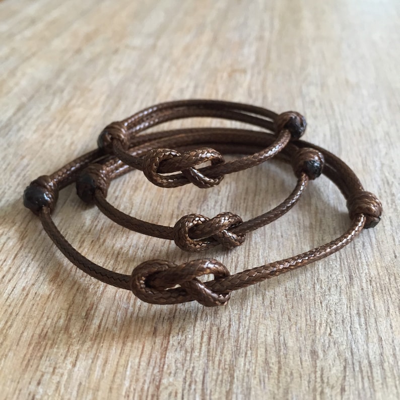 May include: Three brown cord bracelets with knotted ends. The bracelets are stacked on top of each other.